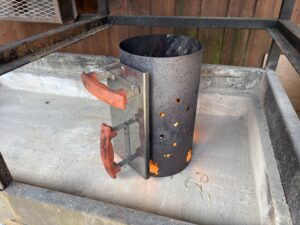 Rusty vertical charcoal chimney starter with orange-handled latch, mounted in a metal grill frame outdoors.