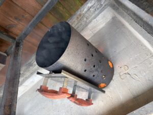 Close-up of a rusted metal smoker or fire pit with vent holes and orange handles, outdoors on a concrete surface.