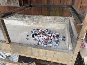 Pile of charcoal briquettes covered in white ash sits in the bottom of a metal barbecue grill with a rusted frame and wooden fence in the background, outdoors.