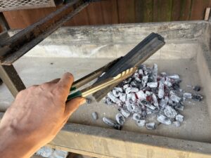 Person holding a metal grate piece over a charcoal grill with white-hot coals visible below.